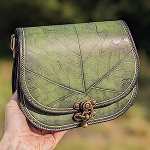 May include: A green leather crossbody bag with a leaf design. The bag has a brass clasp and a strap.