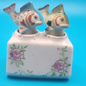 Puede incluir: Un juego de salero y pimentero de cerámica blanca con dos saleros en forma de pez. Los peces están pintados con detalles azules, rojos y verdes. La base está decorada con diseños florales rosas y ribetes dorados.