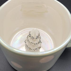 Op de afbeelding: Een witte keramische mok met een parelmoerglans. Binnenin de mok bevindt zich een kleine, witte, boomvormige decoratie met donkere spikkels. De mok heeft een oor en een brede rand.