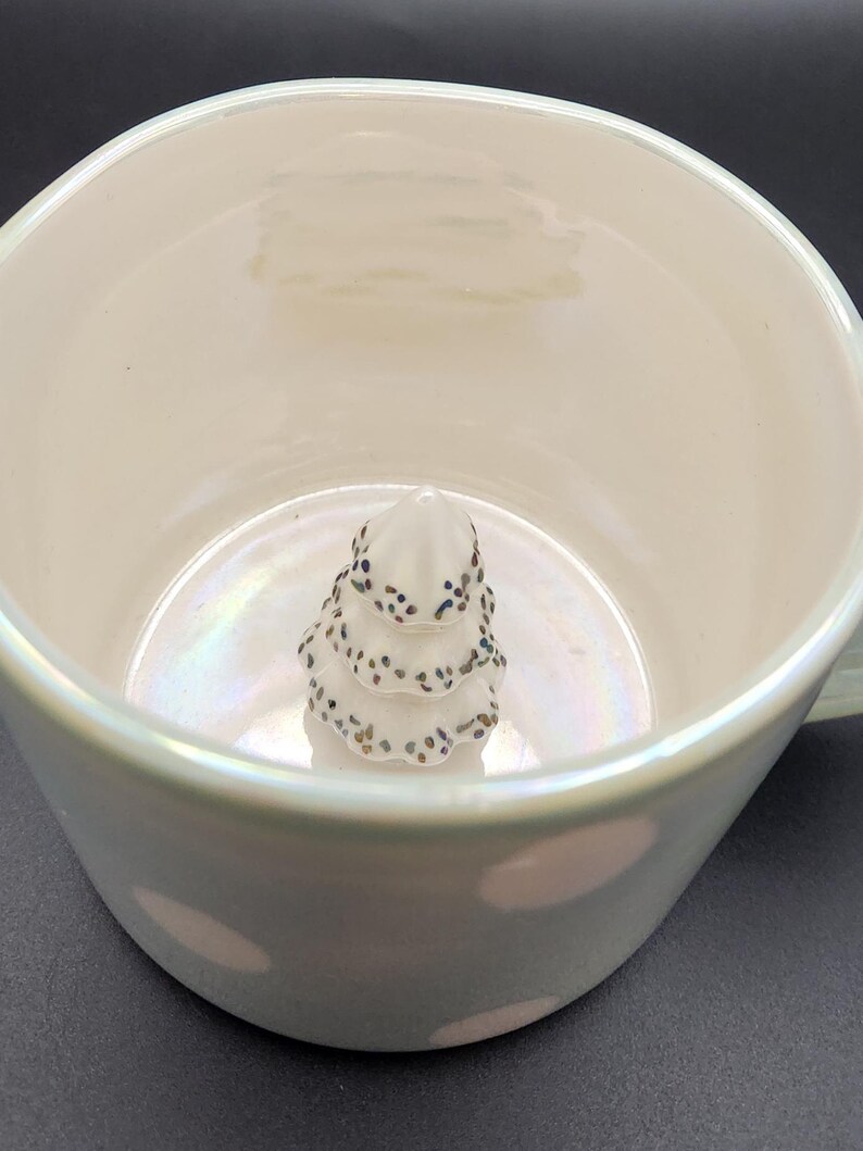 Op de afbeelding: Een witte keramische mok met een parelmoerglans. Binnenin de mok bevindt zich een kleine, witte, boomvormige decoratie met donkere spikkels. De mok heeft een oor en een brede rand.