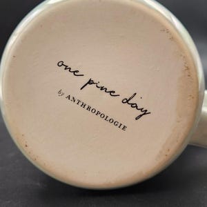 Op de afbeelding: Onderkant van een keramische mok met een lichtgroene rand. De basis is gebroken wit met de woorden "one pine day" en "by ANTHROPOLOGIE" in zwart schrift.