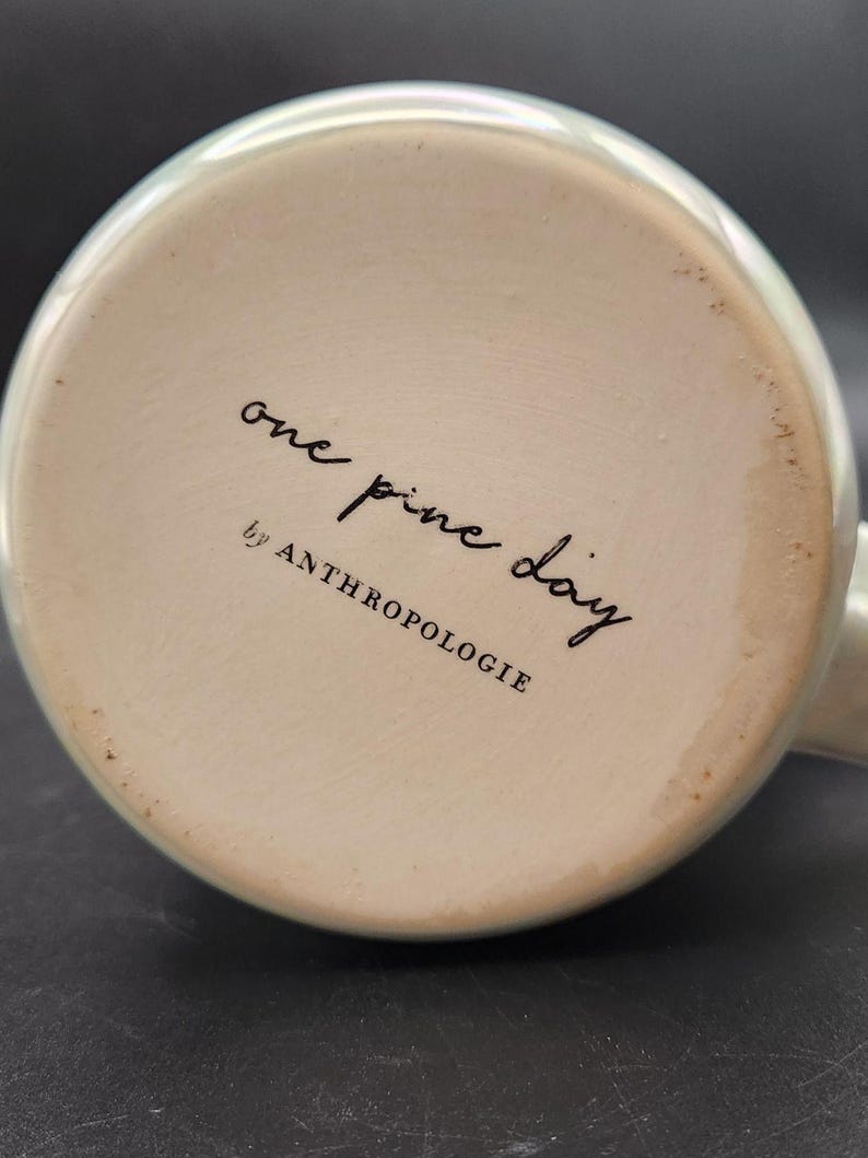 Op de afbeelding: Onderkant van een keramische mok met een lichtgroene rand. De basis is gebroken wit met de woorden "one pine day" en "by ANTHROPOLOGIE" in zwart schrift.