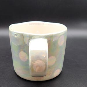 Op de afbeelding: Iridescent keramische mok met een parelmoer afwerking en een witte handgreep. De mok heeft een patroon van zachte roze stippen. De mok is ontworpen voor warme dranken en heeft een brede, comfortabele handgreep.