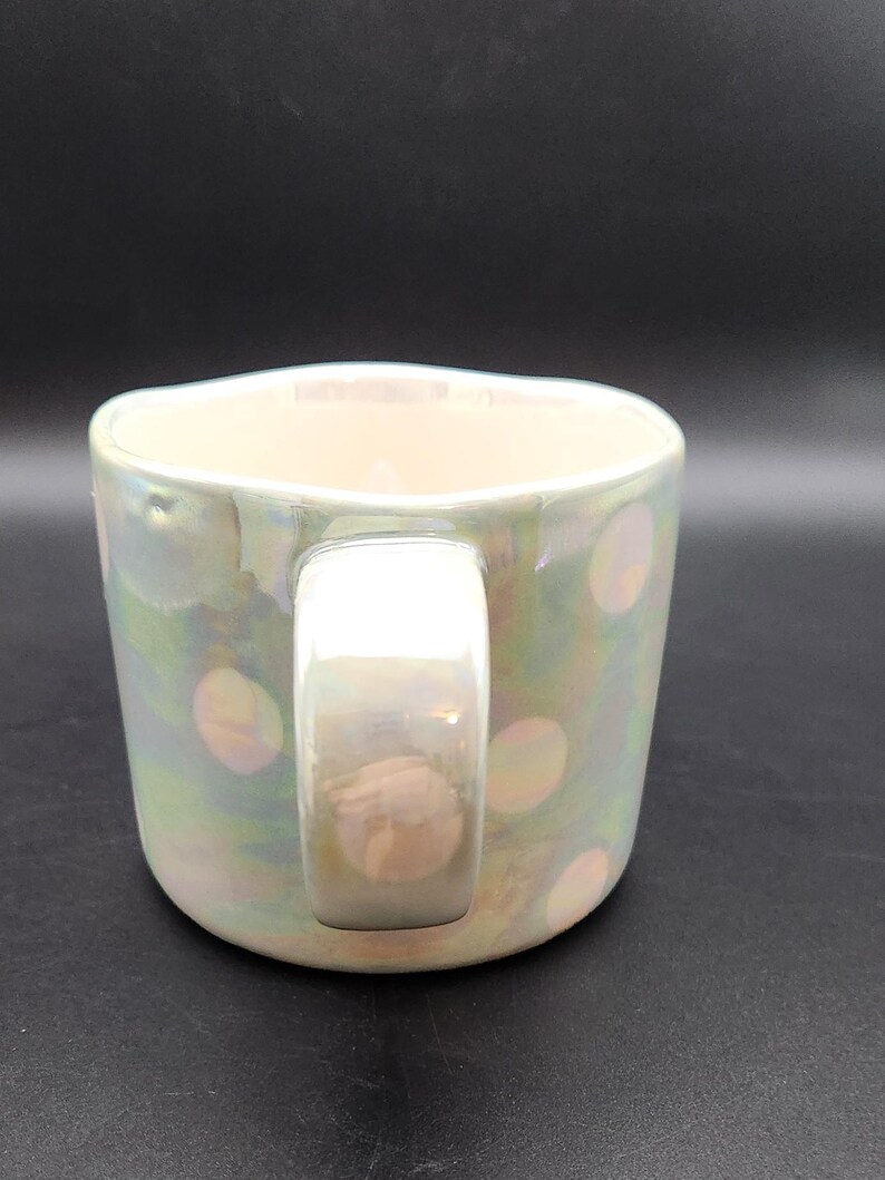 Op de afbeelding: Iridescent keramische mok met een parelmoer afwerking en een witte handgreep. De mok heeft een patroon van zachte roze stippen. De mok is ontworpen voor warme dranken en heeft een brede, comfortabele handgreep.