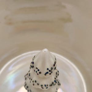Op de afbeelding: Een kleine, witte keramische kerstboom met donkerblauwe en bruine spikkels. De boom staat in een witte kom met iriserende highlights. De afbeelding is een close-up.