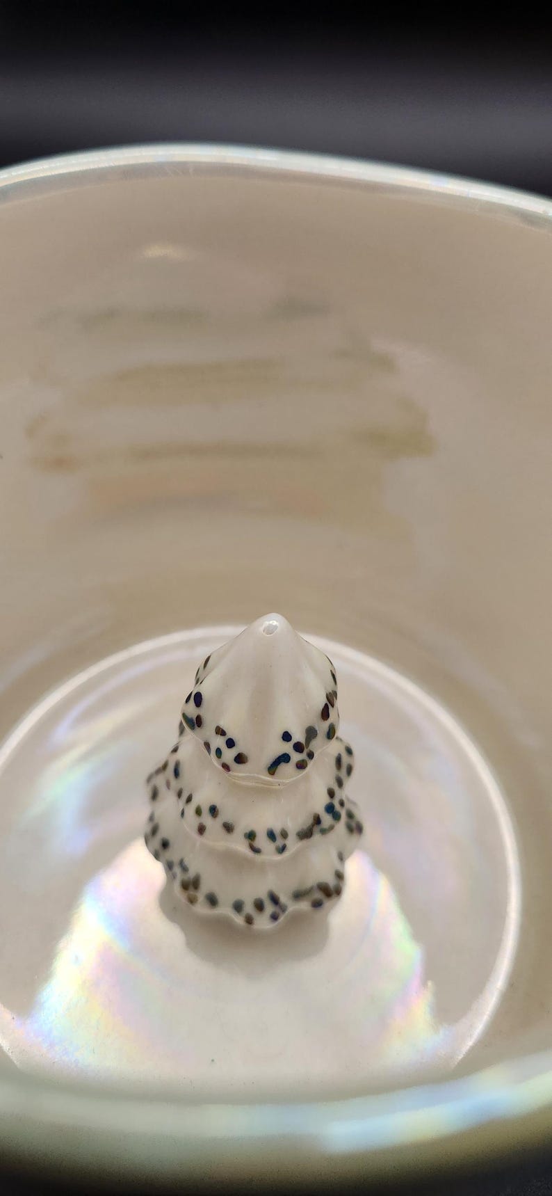 Op de afbeelding: Een kleine, witte keramische kerstboom met donkerblauwe en bruine spikkels. De boom staat in een witte kom met iriserende highlights. De afbeelding is een close-up.