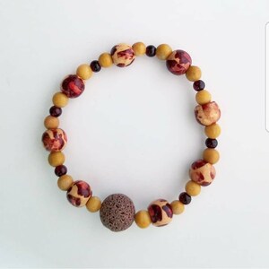 以下が含まれることがあります： 赤と白のパターンが施された、茶色と黄色のビーズで作られたブレスレット。ダークブラウンの溶岩ビーズが1つ付いている。