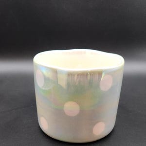 Op de afbeelding: Iridescent keramische beker met een parelmoer afwerking en roze stippen. De beker heeft een afgeronde vorm en een licht golvende rand. Het oppervlak reflecteert een reeks kleuren, waaronder groen en blauw, waardoor een glinsterend effect ontstaat. De beker staat tegen een donkere achtergrond.