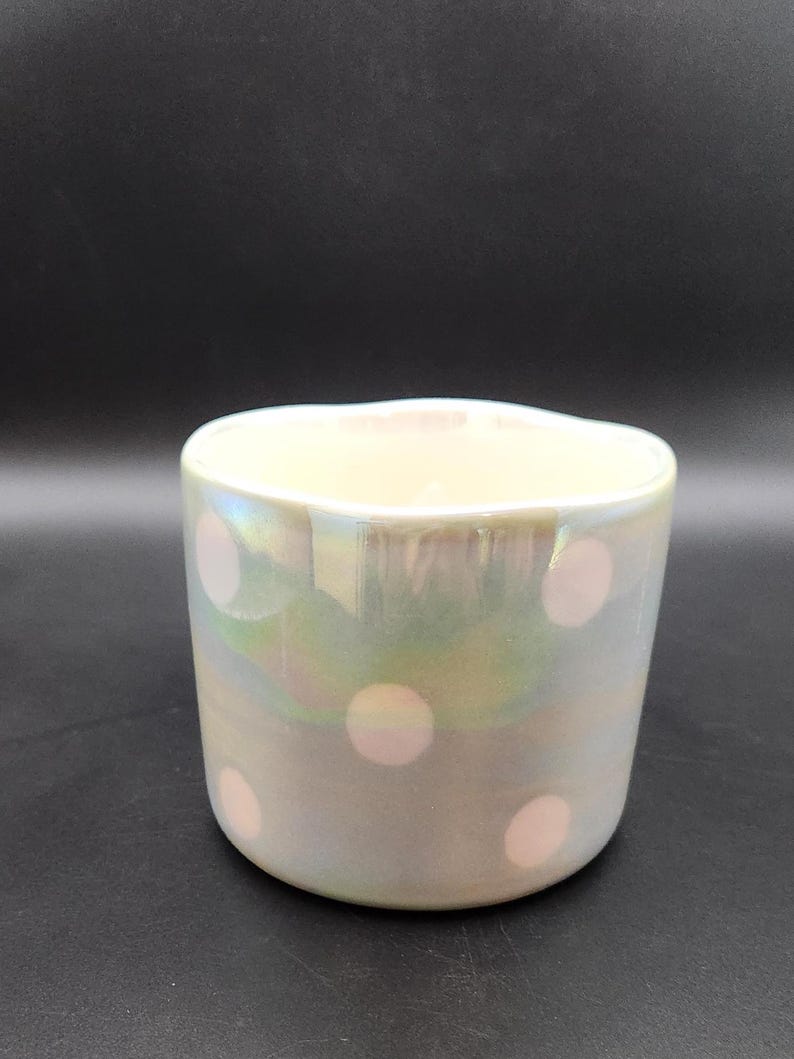Op de afbeelding: Iridescent keramische beker met een parelmoer afwerking en roze stippen. De beker heeft een afgeronde vorm en een licht golvende rand. Het oppervlak reflecteert een reeks kleuren, waaronder groen en blauw, waardoor een glinsterend effect ontstaat. De beker staat tegen een donkere achtergrond.