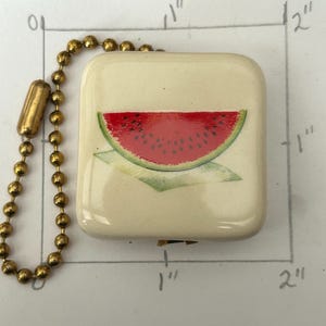 May include: A rectangular, cream-coloured keychain charm featuring a painted watermelon slice. The watermelon has a red interior with black seeds and a green rind. The charm is attached to a gold-coloured ball chain.