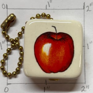 May include: A square keychain with a cream background, featuring a red apple illustration with a white highlight and black stem. The keychain is attached to a gold-coloured ball chain.
