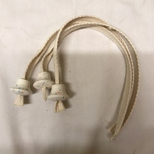 Op de afbeelding: Drie witte keramische olielamp lonten met klokvormige toppen, besprenkeld met kleurrijke verf, verbonden met gebroken witte geweven koorden. De lonten zijn gerangschikt in een gebogen patroon tegen een witte achtergrond. Voor olielampen.