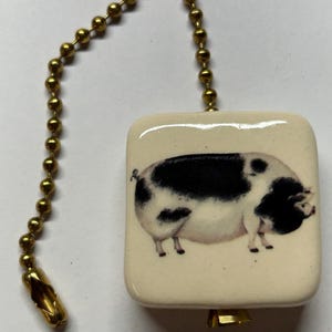 May include: A square, cream-coloured ceramic ceiling fan pull chain ornament featuring a detailed illustration of a black and white spotted pig. The ornament is attached to a metallic gold-toned ball chain with a matching gold-toned connector.