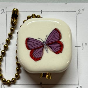 Op de afbeelding: Een vierkante, crèmekleurige plafondventilator trekker met een paars en rood vlinderontwerp. Het is bevestigd aan een goudkleurige ketting. De trekker meet ongeveer 2,5 cm bij 2,5 cm.