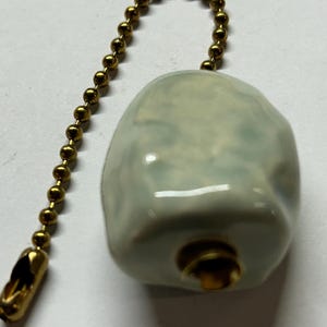 May include: A pale blue ceramic light pull with a gold-coloured chain. The pull has a rounded, irregular shape and a glossy finish. The chain is made of small, round beads and has a decorative end.