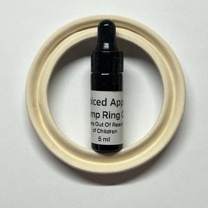 May include: A small black 5 ml dropper bottle with a white label reading Spiced Apple, Pump Ring, Keep Out Of Reach of Children, and 5 ml, positioned inside a circular, off-white, matte-finished ring component with a raised inner edge.