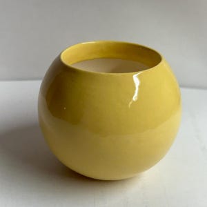 May include: A round, yellow ceramic vase with a glossy finish. The vase has a wide opening and a smooth, spherical shape. The interior is a light beige color. The vase is sitting on a white surface.