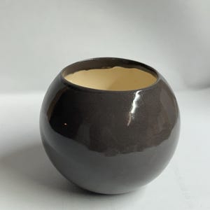 Puede incluir: Un jarrón de cerámica marrón oscuro brillante con forma esférica. El interior es de color beige claro, contrastando con el exterior. El jarrón tiene una abertura ancha y una superficie lisa y reflectante.