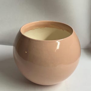 May include: A round, peach-colored ceramic vase with a glossy finish. The interior is a light cream color. The vase has a wide opening and a smooth, curved shape. It is suitable for holding flowers or as a decorative piece.