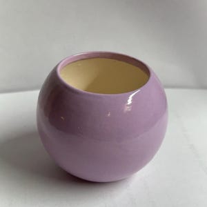 May include: A small, round, lavender-colored ceramic bowl with a glossy finish. The interior is a light yellow color. The bowl has a smooth, spherical shape and is likely used for decorative purposes or as a small container.