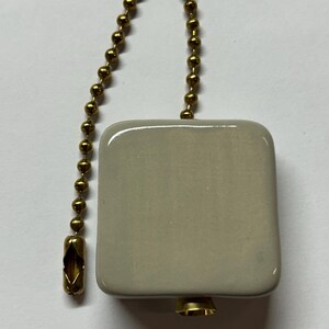 May include: A square, light beige ceramic ceiling fan pull chain with a gold-coloured metal chain and connector. The pull chain is designed to operate a ceiling fan or light fixture.