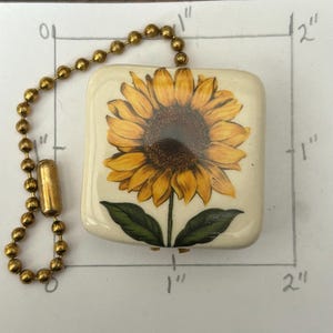 Könnte beinhalten: Ein quadratischer, cremefarbener Keramik-Lichtschalter mit einer detaillierten Sonnenblumenillustration. Die Sonnenblume hat gelbe Blütenblätter und eine braune Mitte mit grünen Blättern. Er ist an einer goldfarbenen Kugelkette befestigt.