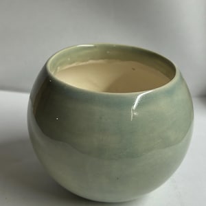 May include: A round, ceramic bowl with a smooth, glossy finish. The exterior is a muted, pale green, while the interior is a creamy off-white. The bowl has a wide opening and a rounded shape, suitable for various uses.