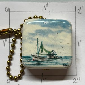 May include: A square, ceramic ceiling fan pull featuring a painted seascape with a sailboat. The artwork depicts a boat with green sails on blue water under a cloudy sky. The pull is approximately 2.5 cm by 2.5 cm and is attached to a gold-coloured ball chain.