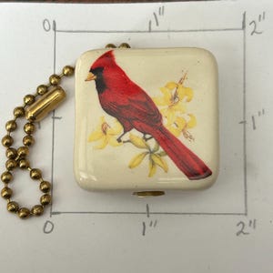 May include: A square, cream-colored trinket box with a detailed image of a red cardinal bird perched on a yellow flower branch. The box is approximately 1 inch by 1 inch and has a gold-colored ball chain attached.
