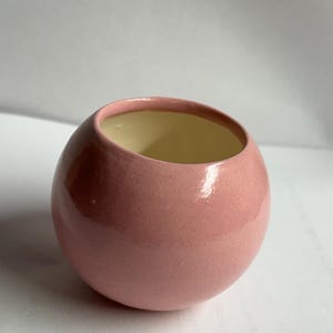 May include: A small, pink ceramic bowl with a glossy finish. The bowl has a rounded shape with a slightly angled opening, revealing a light yellow interior. The smooth surface reflects light, highlighting its spherical form.