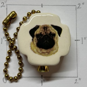 May include: A cream ceramic light pull with a pug design. The pug has a black muzzle and ears, with tan fur. It's attached to a gold-coloured chain and connector. The light pull is approximately 5 cm in size.
