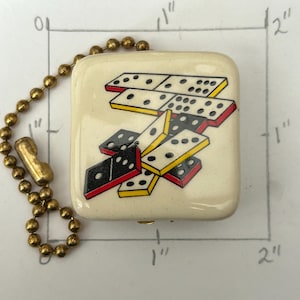 May include: A square, off-white keychain with a gold-colored ball chain. The keychain features a colorful illustration of dominoes with black, red, and yellow accents. The keychain is approximately 1 inch by 1 inch.