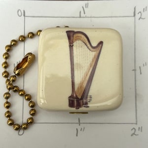 May include: A square keychain with a cream-colored background features a detailed illustration of a harp in shades of brown and gold. The keychain is attached to a gold-colored ball chain.