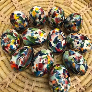 Puede incluir: Una colección de once huevos de cerámica con un diseño moteado y colorido. Los huevos son blancos con un patrón de salpicaduras de pintura roja, azul, verde y amarilla. Están dispuestos en una cesta tejida.