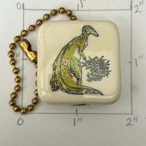 May include: A square, off-white keychain with a gold-colored chain. The keychain features a green and yellow dinosaur illustration with a black outline. The dinosaur is standing near a cluster of black plants.