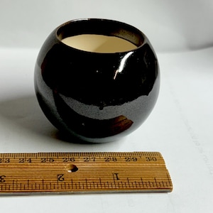 Puede incluir: Un pequeño cuenco de cerámica marrón oscuro brillante, de forma redondeada. El interior es de color crema claro. Una regla de madera se coloca junto al cuenco, mostrando las medidas en centímetros.