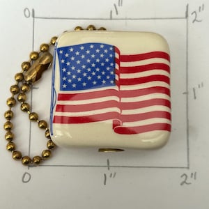 Könnte beinhalten: Ein quadratischer Schlüsselanhänger, ca. 2,5 cm x 2,5 cm, mit dem Design der amerikanischen Flagge. Die Flagge hat rote und weiße Streifen und ein blaues Feld mit weißen Sternen. Er ist an einer goldfarbenen Kugelkette befestigt.