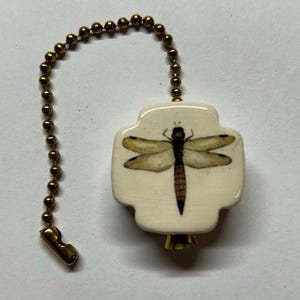 May include: A cream-colored ceramic ceiling fan pull chain ornament featuring a cross-shaped design with a detailed dragonfly illustration in shades of brown and yellow, attached to a metallic gold-toned beaded chain with a connector.