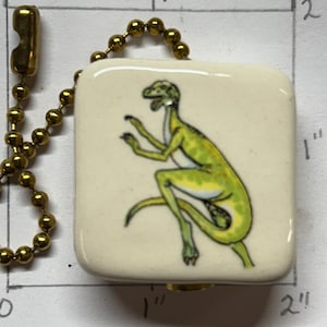 以下が含まれることがあります： 金色のチェーンが付いた、正方形のオフホワイトのペンダント。ペンダントには、緑と黄色のトカゲのイラストが描かれています。ペンダントのサイズは約2.5cm x 2.5cmです。