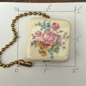 May include: A square, off-white ceramic pull chain pendant with a floral design. The pendant features a pink rose and other colorful flowers. It measures approximately 1 inch by 1 inch and is attached to a gold-colored ball chain.
