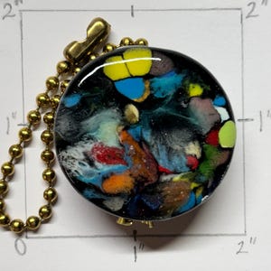 May include: A round, abstract art pendant with a black background and colorful splashes of blue, yellow, red, and green. The pendant is approximately 2 inches in diameter and attached to a gold-colored ball chain.