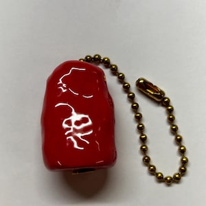 Puede incluir: Un dije rojo brillante de forma rectangular redondeada con un diseño gráfico blanco en la parte frontal. El dije está unido a una cadena de bolas de metal en tono dorado, que incluye una pequeña pieza de conexión para cerrar la cadena.