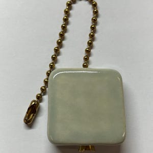 Op de afbeelding: Een vierkante, crèmekleurige keramische lichtschakelaar met een goudkleurige ketting en connector. De schakelaar heeft afgeronde hoeken en een glanzende afwerking. De ketting is gemaakt van kleine, ronde kralen. De connector heeft een decoratieve, langwerpige vorm.