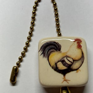May include: A square, cream-colored ceramic ceiling fan pull chain ornament featuring a detailed illustration of a rooster with black, yellow, and red plumage. The ornament is attached to a gold-toned metal ball chain with a matching connector.