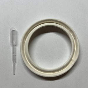 Op de afbeelding: Een doorzichtige plastic pipet naast een cirkelvormige, gebroken witte siliconen pakking. De pakking heeft een verhoogde buitenrand en een verzonken binnenkanaal, terwijl de pipet een bolvormige bovenkant en een smalle, langwerpige steel heeft.