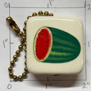 以下が含まれることがあります： 正方形のオフホワイトのチャーム。スイカのスライスが描かれています。スイカは赤い内部に黒い種と緑色の皮があります。金色のボールチェーンと留め金が付いています。チャームのサイズは約2.5cm x 2.5cmです。