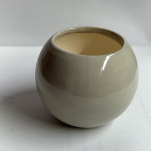 May include: A small, round, ceramic vase with a smooth, glossy, gray exterior. The interior is a light beige color. The vase has a wide opening and a spherical shape, suitable for small plants or decorative use.