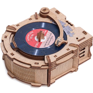 Könnte beinhalten: Ein Holzmodell eines Plattenspielers aus ineinandergreifenden Puzzleteilen. Die mittlere Scheibe hat ein rot-weißes Etikett mit dem Text Adventure Music, Stereo und Song of Puzzles. Ein Tonarm ruht auf der dunkelblauen, kreisförmigen Scheibe.