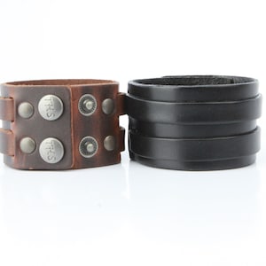 K&ouml;nnte beinhalten: Zwei Lederarmb&auml;nder. Eines ist braun mit silberfarbenen Nieten und dem Text "THS" auf den Druckkn&ouml;pfen. Das andere ist schwarz mit einem geschichteten Design. Beide Manschetten sind breit und haben ein robustes Aussehen.