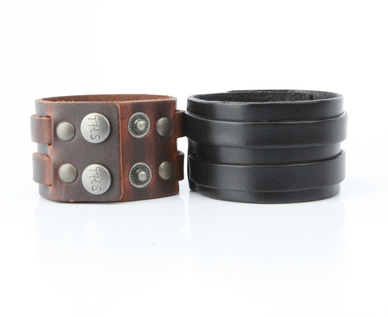 K&ouml;nnte beinhalten: Zwei Lederarmb&auml;nder. Eines ist braun mit silberfarbenen Nieten und dem Text "THS" auf den Druckkn&ouml;pfen. Das andere ist schwarz mit einem geschichteten Design. Beide Manschetten sind breit und haben ein robustes Aussehen.